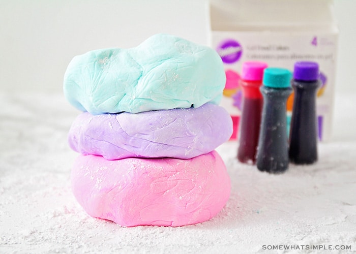 three flattened balls of marshmallow fondant stacked on top of each other on a counter covered with powdered sugar. The ball of fondant on top is light blue, the middle one is light purple and the bottom one is light pink. In the background are three bottles of Wilton gel food coloring.