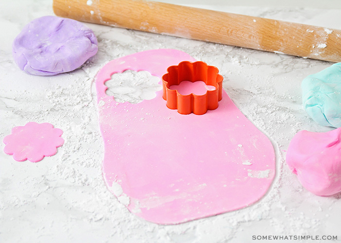 on a counter dusted with powdered sugar a piece of light pink marshmallow fondant has been rolled out flat and a flower shaped piece has been cut out near the top. Above it is a flattened ball of homemade purple marshmallow fondant and a wood rolling pin.