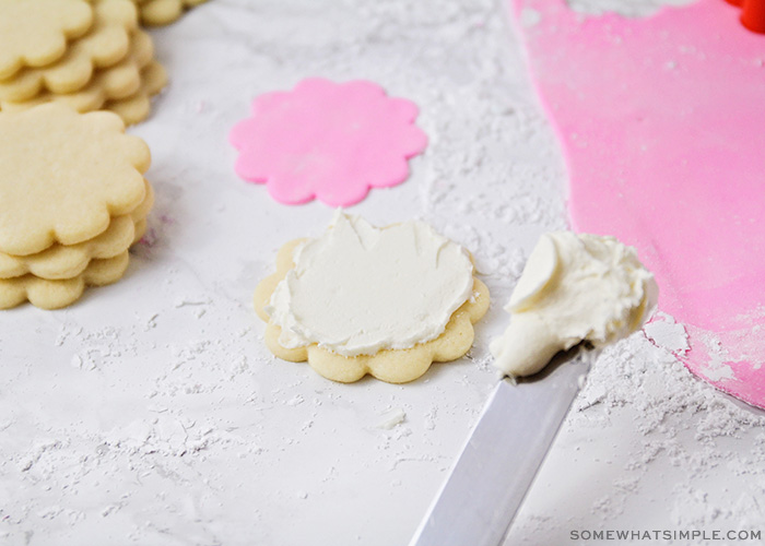 a sugar cookie with white fondant made from marshmallows is on a counter. Hoovering over the air is an icing spatula with a glob of fondant on the tip. Next to the cookie are stacks of additional unfrosted flower-shaped sugar cookies. On the other side of the image is flattened pink marshmallow fondant.