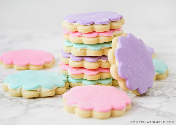 a stack of sugar cookies topped with either pink, light green or purple marshmallow fondant. One cookie with purple fondant is on it's side leaning against the stack of cookies and a few other cookies are laying flat on the counter around the stack.