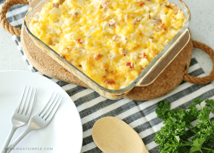 a square casserole dish filled with ham and potato casserole topped with melted cheese sitting on a hot pad. Next to the casserole dish is a white plate with two forks on top. Next to the plate is a wooden serving spoon and a clove of parsley. Everything is sitting on a black and white striped kitchen towel.