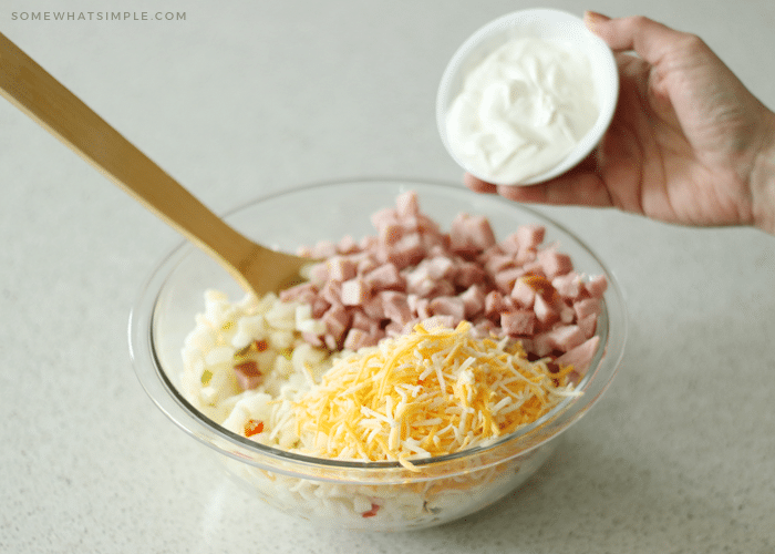 a glass bowl with a wood spoon inside that's filled with shredded cheese, diced ham and potatoes with a hand about to pour a small bowl of sour cream into the larger bowl.