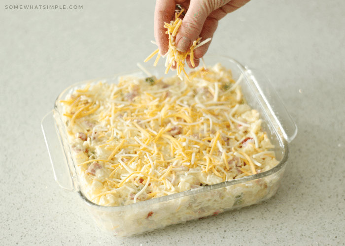 a hand sprinkling shredded cheese over the top of a casserole dish filled with ham and potato casserole