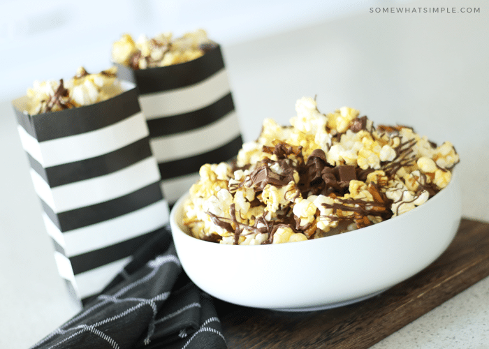 a white bowl filled with gourmet popcorn with drizzled chocolate on top mixed with pieces of kit kat bars and pretzels. Behind the bowl are two black and white striped bags filled with more chocolate covered popcorn