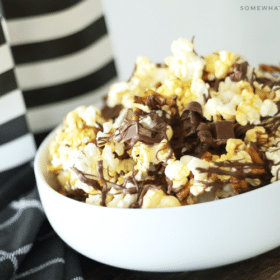 a white bowl filled with gourmet popcorn with drizzled chocolate on top mixed with pieces of kit kat bars and pretzels. Behind the bowl are two black and white striped bags filled with more chocolate covered popcorn