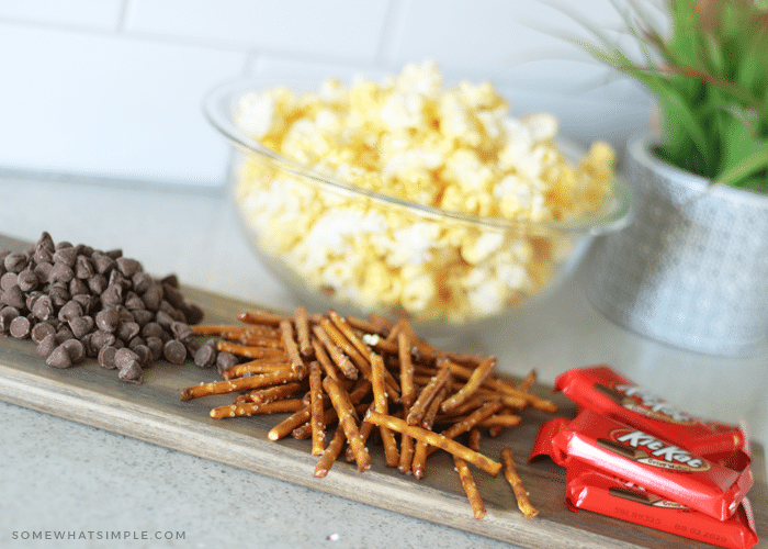 a bowl of popped popcorn sitting on the counter behind piles of chocolate chips, pretzel sticks and kit kat bars which are the ingredients for this gourmet chocolate popcorn