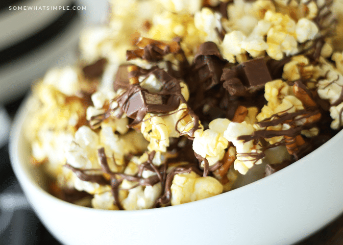 a close up of a white bowl of gourmet popcorn mixed with small pieces of kit kat bars and pretzels with drizzled chocolate over the top that was made using this easy recipe