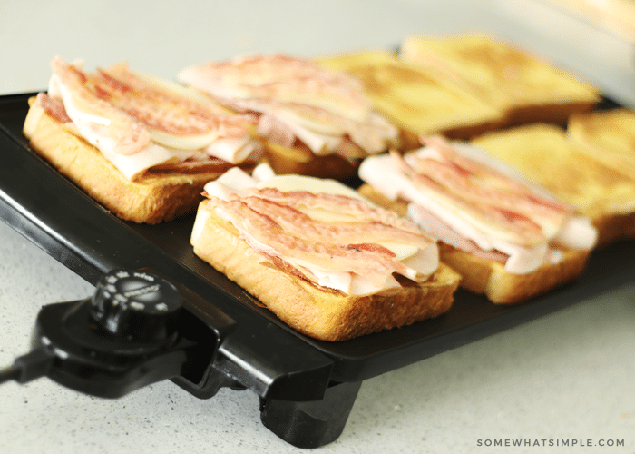 slices of bread cooking on top of a black griddle. The bread on the left side of the griddle are topped with ham, turkey and bacon, while the slices on the right side don't have anything on top of them.