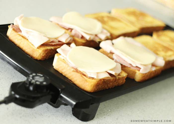 slices of bread cooking on top of a black griddle. The bread on the left side of the griddle are topped with ham, turkey and white cheese, while the slices on the right side are golden brown but don't have anything on top of them.