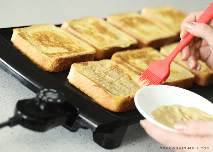 a hand brushing Dijon mustard on a slice of bread that is cooking on a griddle. There are 7 other slices baking that are golden brown but have not been topped with anything.