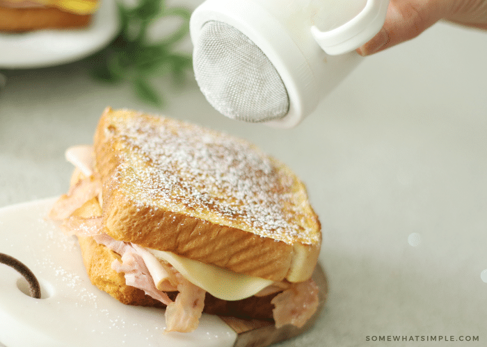a hand sprinkling powdered sugar on top of a cooked Monte Cristo sandwich.