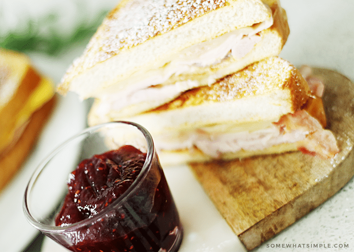 a Monte Cristo sandwich that has been cut in half and the two slices are stacked on top of each other on a wood cutting board. Next to the cutting board is a small glass cup filled with jam.