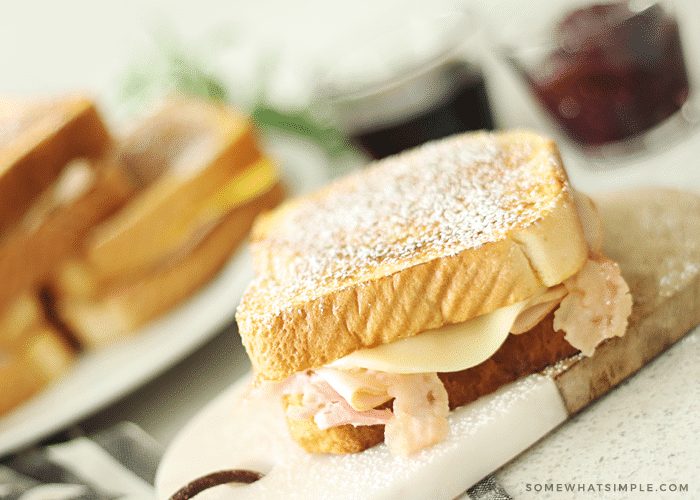 a monte cristo sandwich made using this Disneyland copycat recipe, on a cutting board topped with powdered sugar. Cheese, ham and turkey are piled high and falling out of the side of the sandwich. In the background there's a white plate of additional sandwiches. Next to the plate are two small dipping cups; one filled with maple syrup and the other with jam.