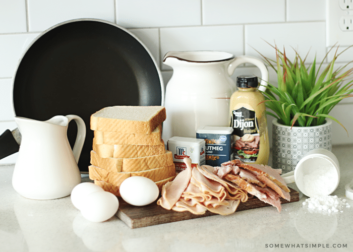 The ingredients for making a Monte Cristo; eggs, a stack of bread, ham, turkey, nutmeg, cinnamon, powdered sugar and Dijon mustard, on a counter.