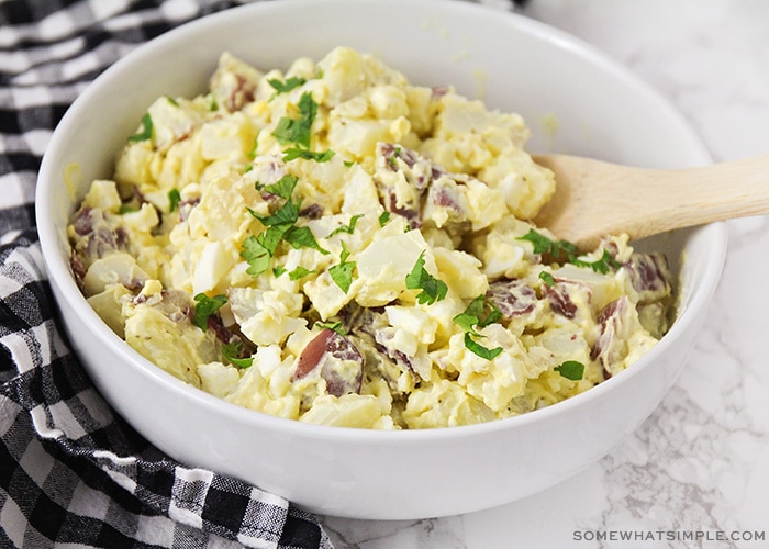 a white bowl filled with this easy potato salad recipe and topped with parsley with a wooden spoon in the bowl.