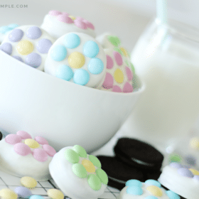 a white bowl filled with chocolate covered Oreos that have pastel colored m&ms on them. On the counter next to the bowl are more chocolate covered oreos and regular oreos with a pitcher of milk in the background.