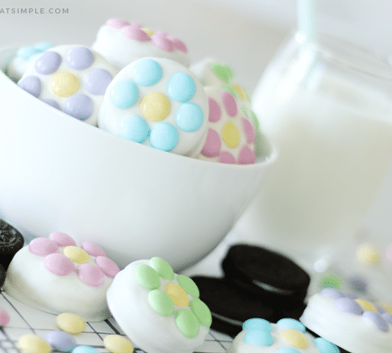 a white bowl filled with chocolate covered Oreos that have pastel colored m&ms on them. On the counter next to the bowl are more chocolate covered oreos and regular oreos with a pitcher of milk in the background.