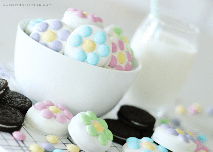 a white bowl filled with chocolate covered Oreos that have pastel colored m&ms on them. On the counter next to the bowl are more chocolate covered oreos and regular oreos with a pitcher of milk in the background.