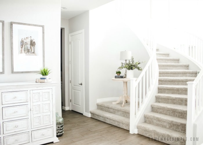 the entry way of a home with a winding staircase. A small wooden table is on an elevated landing. There is also a large white cabinet along the wall in the entry way.