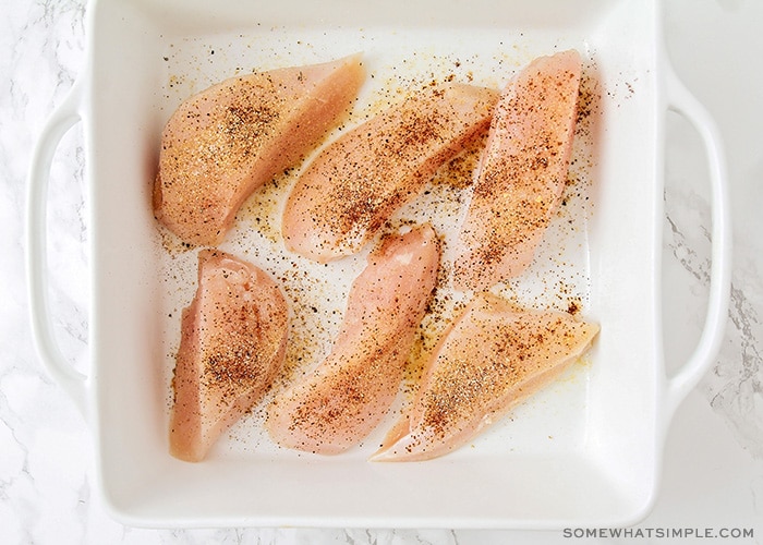 six raw chicken breasts in a white baking dish with seasoning sprinkled over the top