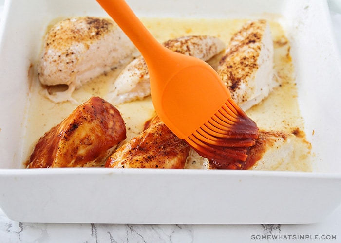an orange silicone basting brush that is basting bbq sauce over the baked breasts. The three breasts in the front have been basted with sauce but the other three have not.