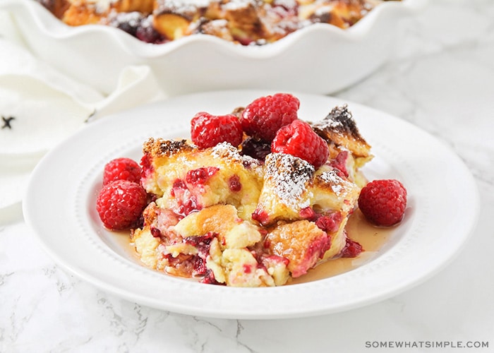 a white plate filled with baked raspberry french toast that is topped with fresh whole raspberries and syrup