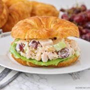 a chicken salad sandwich made with a croissant on a white plate. More croissants and a pile of grapes are laying on the counter behind the plate