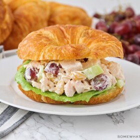 a chicken salad sandwich made with a croissant on a white plate. More croissants and a pile of grapes are laying on the counter behind the plate
