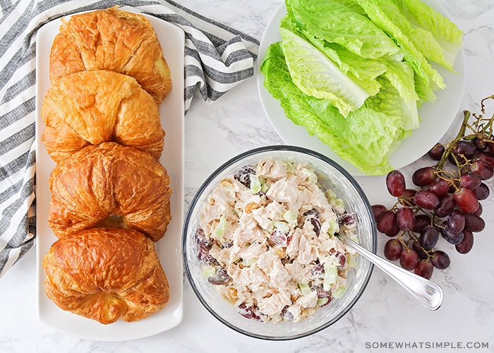 a bowl of chicken salad next to a tray of croissants, grapes and leaves of lettuce 