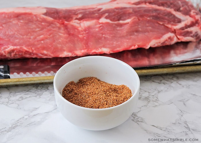 a rack of raw ribs laying on a baking sheet with a small white bowl in front filled with a homemade barbecue dry rub