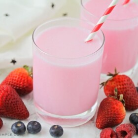 a glass filled with a pink yogurt drink with a striped straw in the cup. Surrounding the glass are several strawberries, blueberries and raspberries.