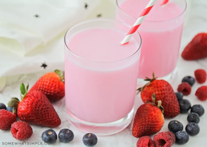 a glass filled with a pink yogurt drink with a striped straw in the cup. Surrounding the glass are several strawberries, blueberries and raspberries.