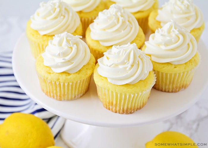 a white cake stand with several lemon cupcakes topped with a cream cheese frosting and white sprinkles. There are a couple of lemons on the counter next to the cake stand.