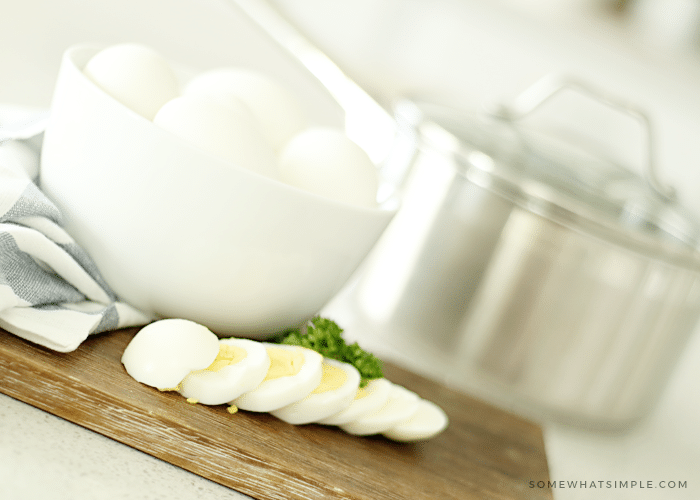 a white bowl filled with hard boiled eggs sitting on a wood cutting board. In front of the bowl is a hard boiled egg that has been sliced into several pieces. Behind the bowl is a stainless steel pot.