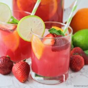 two glasses of non-alcoholic sangria punch that has a light pink color. slices of strawberries and oranges are floating in the cup with a slice of lime sitting on the rim of the glass. Fruit is scattered on the counter around the glass with a pitcher full of punch behind them.