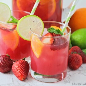 two glasses of non-alcoholic sangria punch that has a light pink color. slices of strawberries and oranges are floating in the cup with a slice of lime sitting on the rim of the glass. Fruit is scattered on the counter around the glass with a pitcher full of punch behind them.