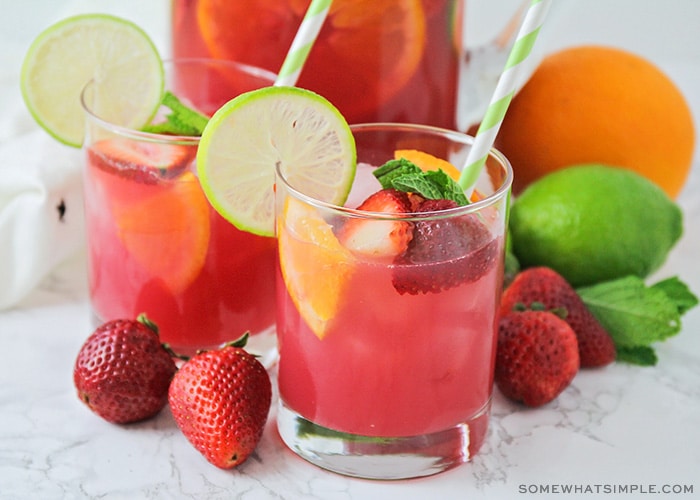 two glasses of non-alcoholic sangria punch that has a light pink color. slices of strawberries and oranges are floating in the cup with a slice of lime sitting on the rim of the glass. Fruit is scattered on the counter around the glass with a pitcher full of punch behind them.