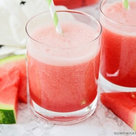 two glasses filled with this watermelon cooler recipe. Each glass has a green and white striped straw and slices of watermelon are next to the cups on the counter