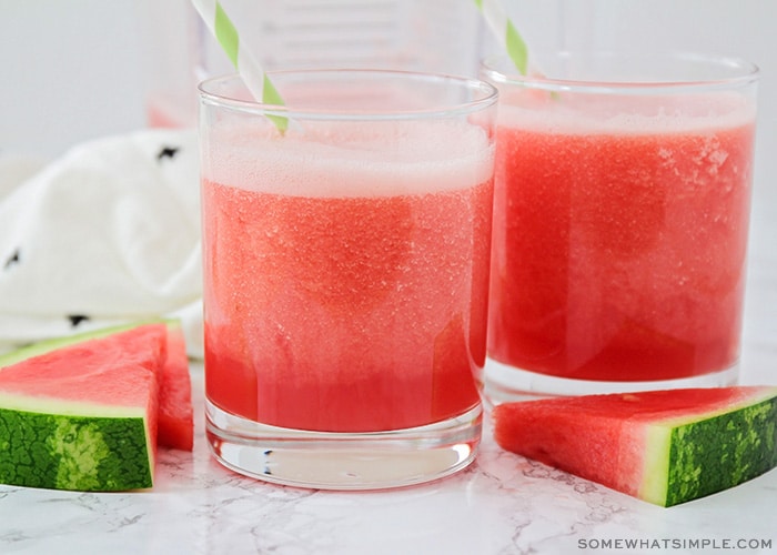 two glass filled with watermelon punch with slices of watermelon laying next to them on the counter