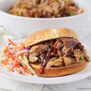 a pulled pork sandwich made in a slow cooker topped with bbq sauce and a side of coleslaw on a white plate.