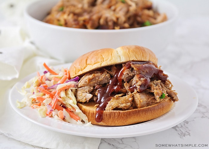 a pulled pork sandwich made in a slow cooker topped with bbq sauce and a side of coleslaw on a white plate.