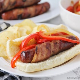 a hot dog wrapped in bacon on a bun, topped with grilled onions and red bell peppers with a side of potato chips on a plate.