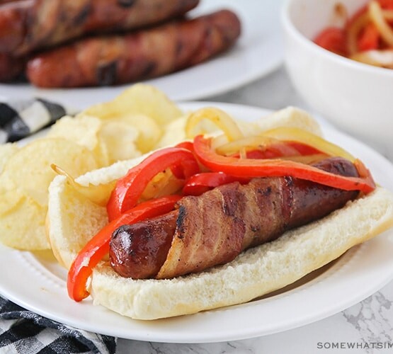 a hot dog wrapped in bacon on a bun, topped with grilled onions and red bell peppers with a side of potato chips on a plate.