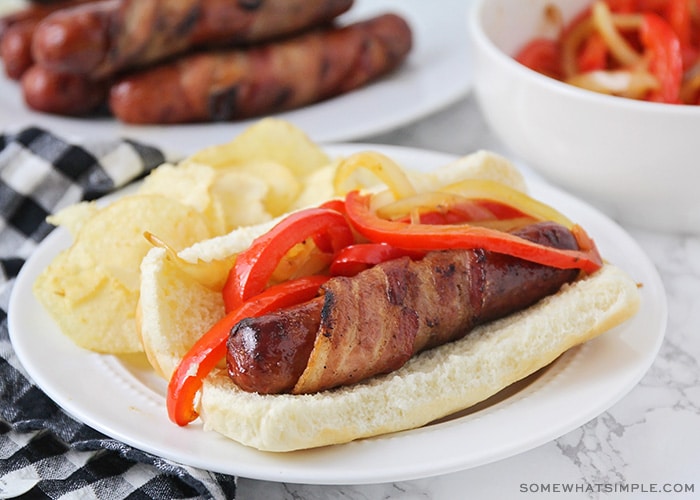 a hot dog wrapped in bacon on a bun, topped with grilled onions and red bell peppers with a side of potato chips on a plate. 