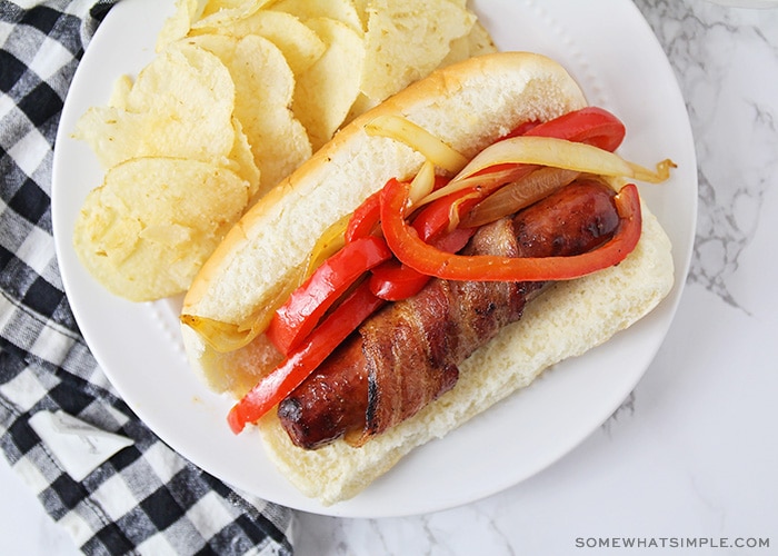 looking down on a hot dog topped with red peppers, grilled onions and bacon next to a side of potato chips.