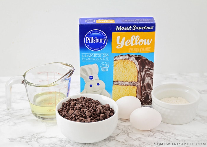 a box of yellow cake mix, a bowl of chocolate chips, two eggs and some oil and flour on a counter
