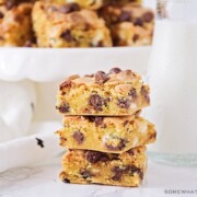 a stack of chocolate chip cookie bar squares made from cake mix next to a jar of milk with a cake stand filled with more bars behind it.