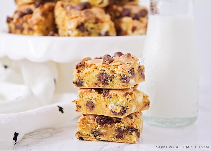 a stack of chocolate chip cookie bar squares made from cake mix next to a jar of milk with a cake stand filled with more bars behind it.