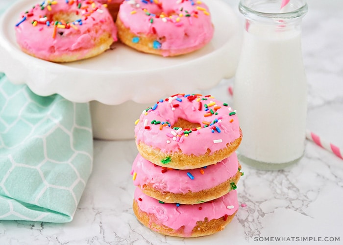 a stack of cake mix donuts topped with a pink glaze and sprinkles next to a cake stand with more donuts and a glass jar of milk