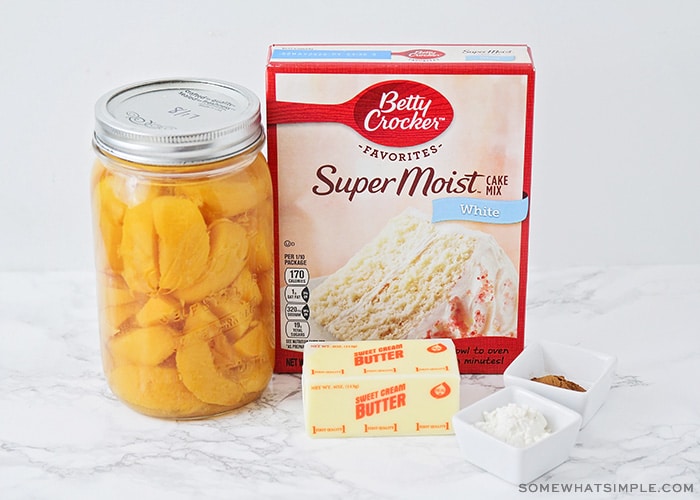 a jar of fresh peaches, a box of white Betty Crocker cake mix, a stick of butter and some cinnamon and flour on the counter